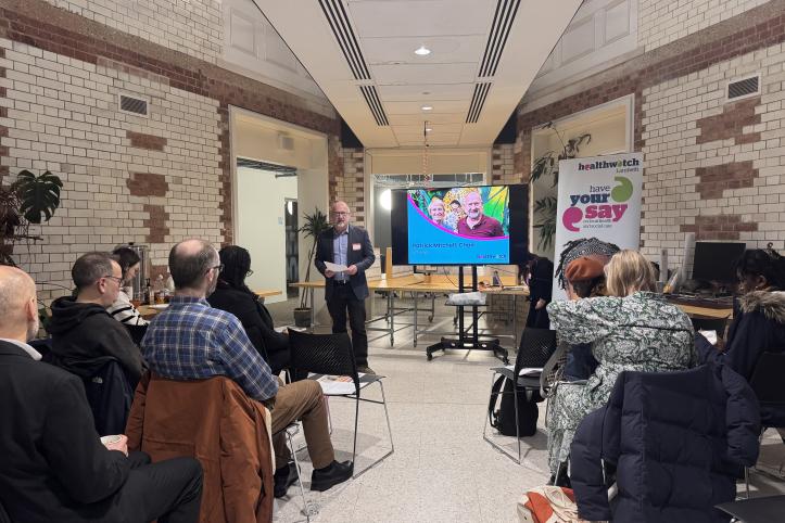 A speaker presenting to a seated audience in a modern, tiled space with a Healthwatch banner visible.