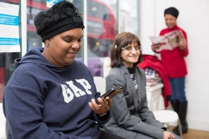 Three women in a bright room; one in a navy hoodie smiles at her phone, another in a gray blazer smiles at the camera, and a third in a red top reads a magazine in the background. A double-decker bus is visible through the window.